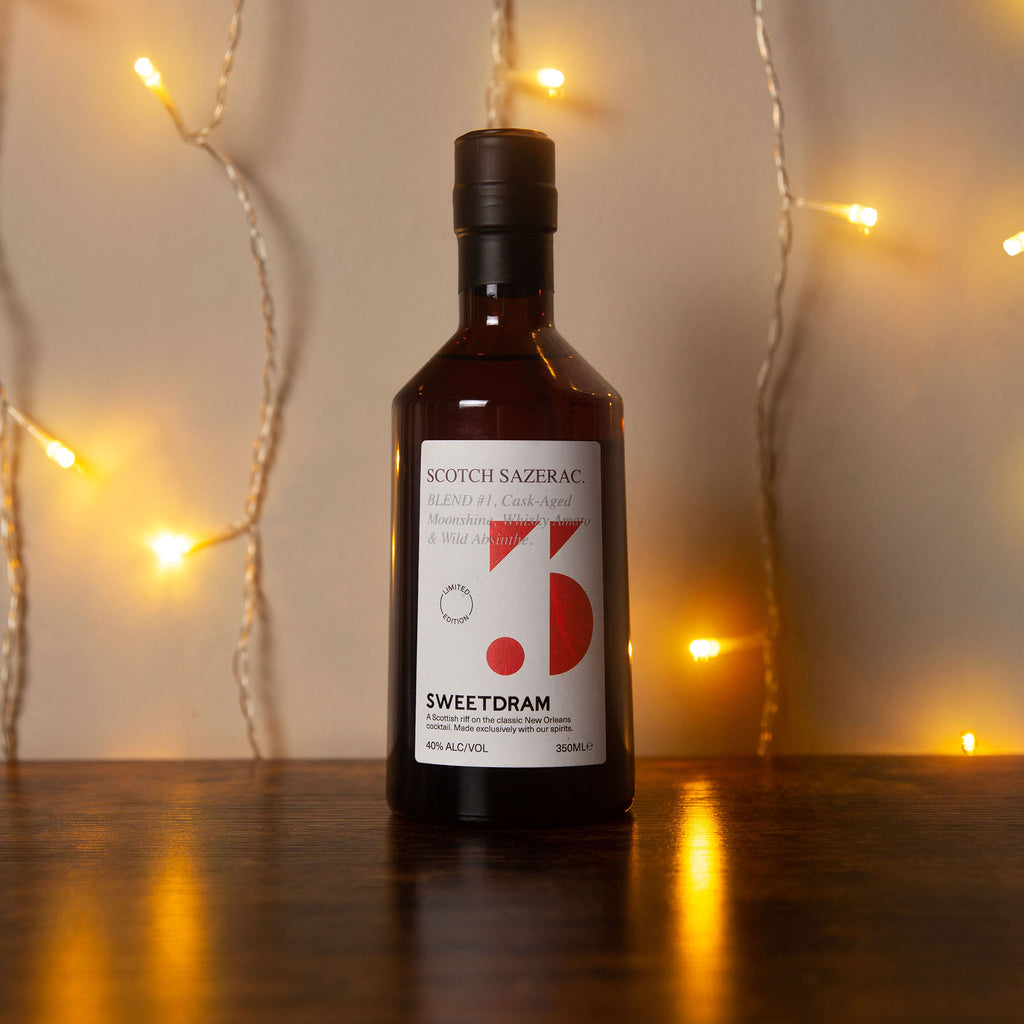 350ml bottle of Scotch Sazerac with red dram logo sitting on a wooden table against a wall of warmly lit fairy lights.