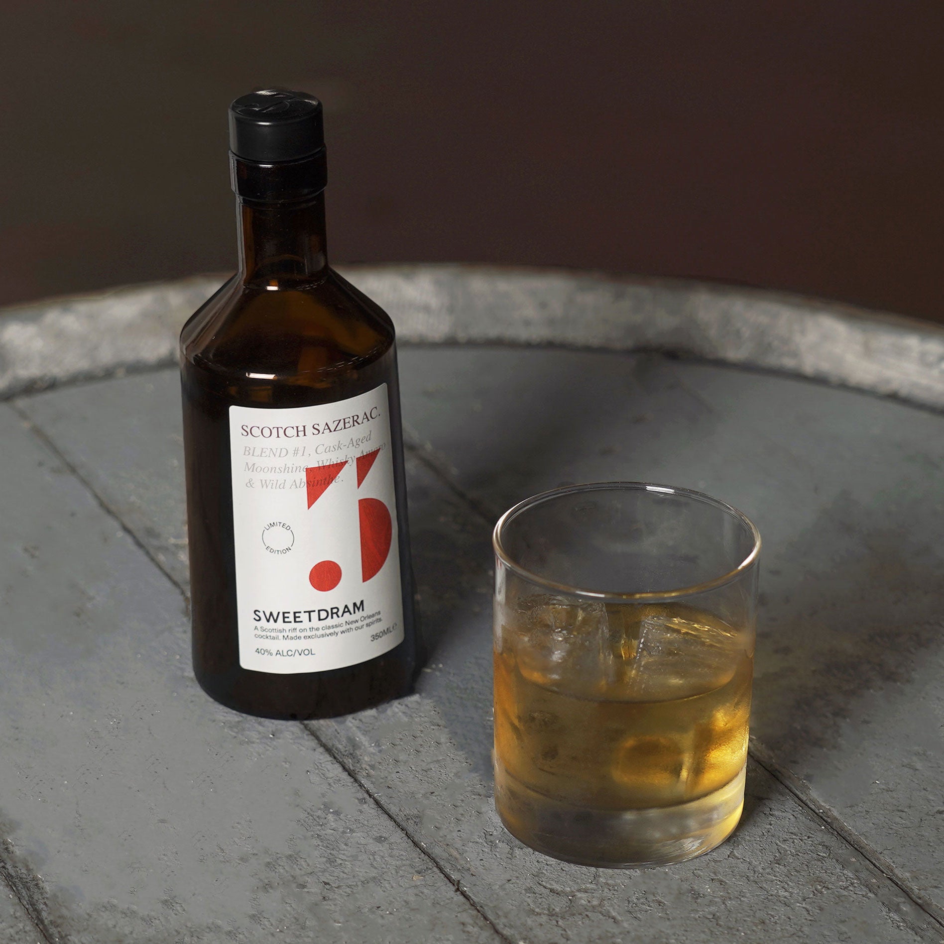 350ml bottle of Scotch Sazerac with red dram logo, sitting on a grey cask with a rocks glass filled with ice and a Sazerac serve.