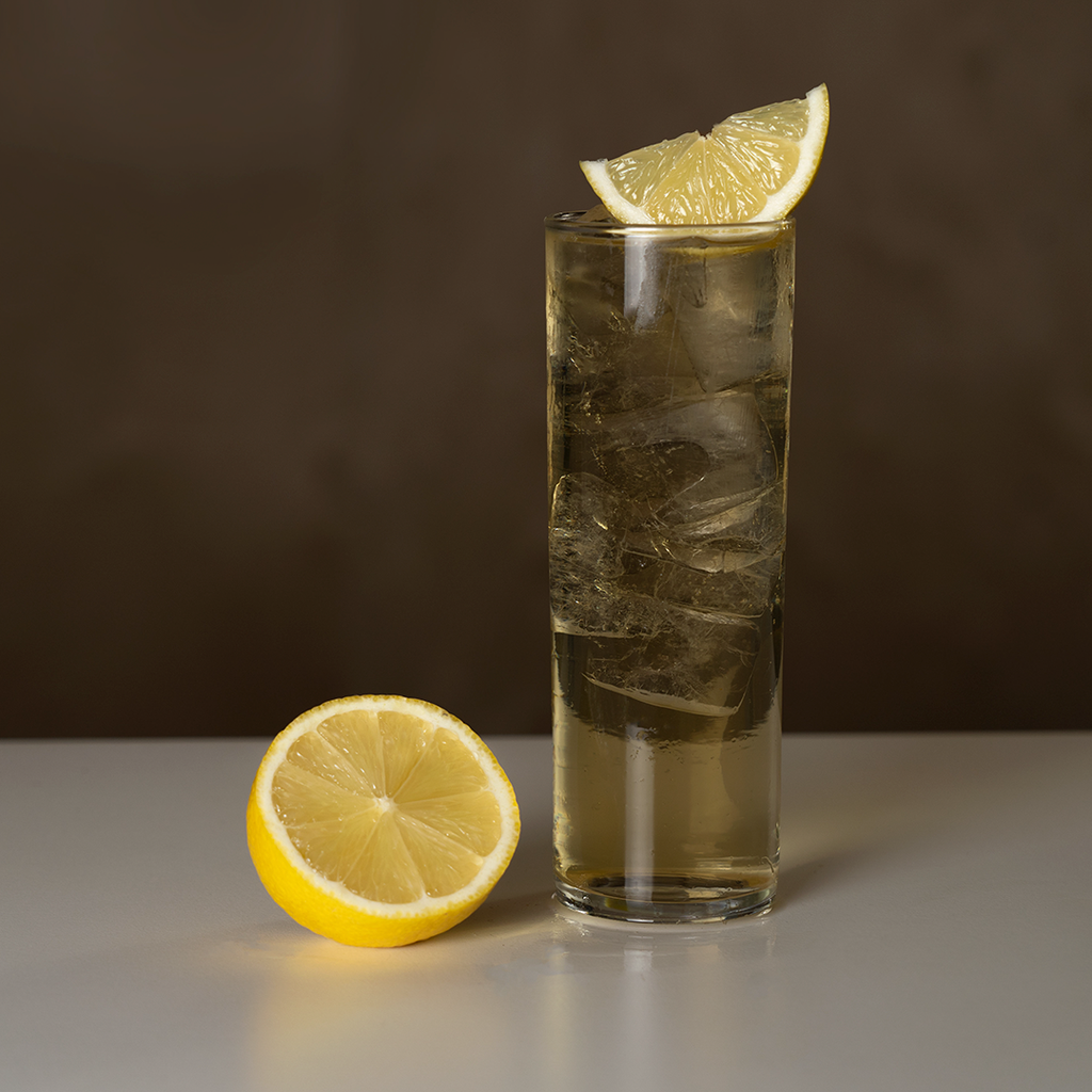 Highball glass with Blend #1 whisky, ginger and ice garnished with lemon wedge. Half lemon sitting next to glass on white table.